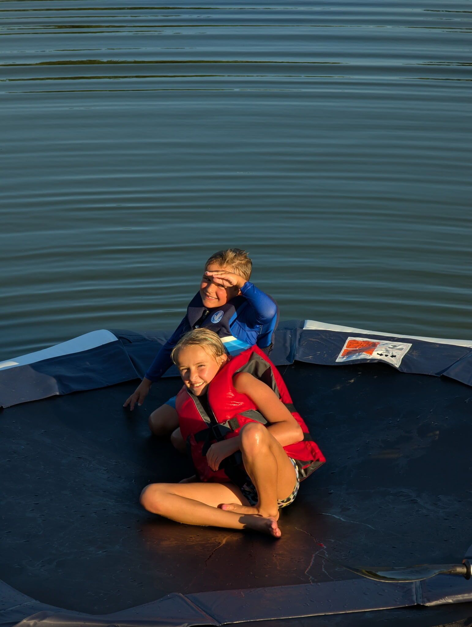 Two people sitting on a trampoline by a body of water.
