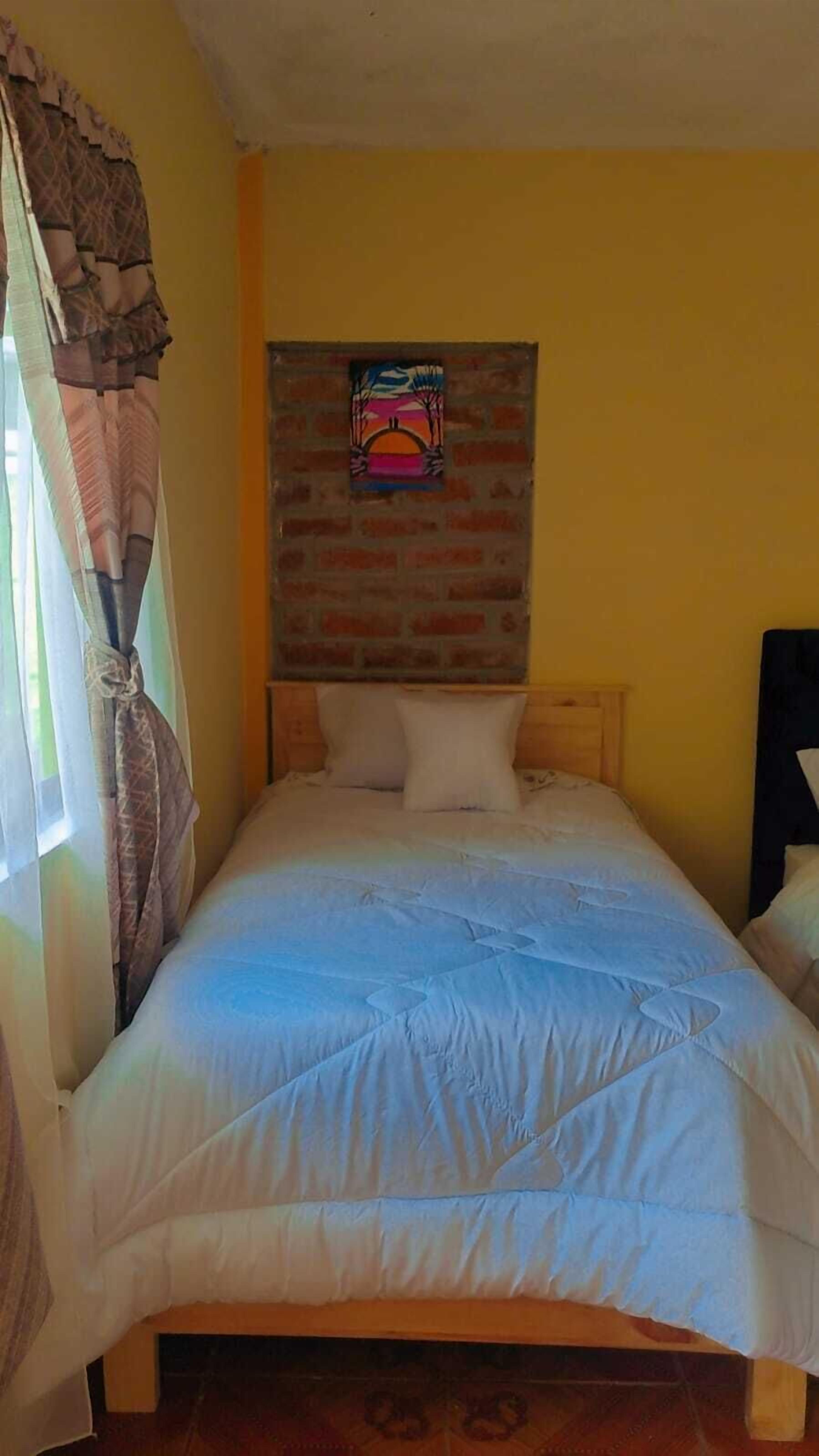A single bed with a wooden headboard, a brick wall, and a small painting above it.