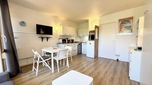 Un espai vital compacte amb una petita cuina, una zona de menjador amb mobles blancs i un televisor fixat a la paret.
