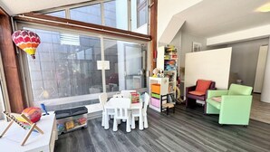 Una habitació amb una finestra gran, una taula amb cadires, una prestatgeria i dues cadires de colors.