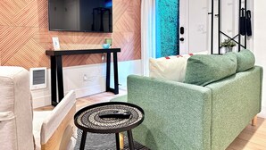 A modern living room with a flat-screen TV mounted on a wooden wall, a green sofa, a small round table with a remote, and a door with a decorative blue panel. - Studio by the Sound - King Bed • Fire Pit • EV Charger • Pet Friendly (Seattle)