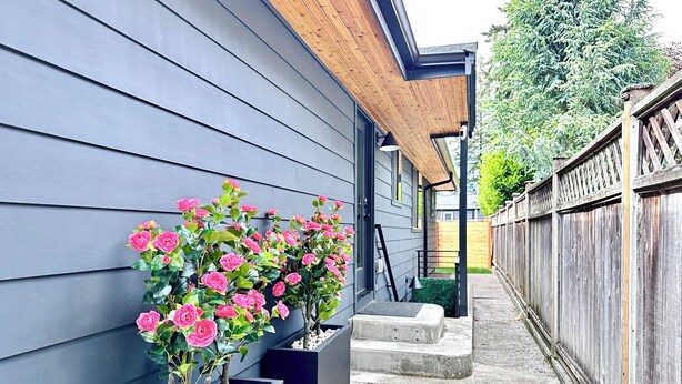 A narrow pathway flanked by a modern building with grey siding and a wooden roof. There are two planters with pink flowers and a wooden fence in the background. - Studio by the Sound - King Bed • Fire Pit • EV Charger • Pet Friendly (Seattle)
