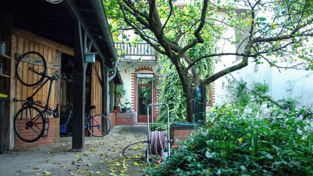 Une ruelle étroite, avec des vélos garés sur le côté gauche, une chaise en métal et un arbre au centre.