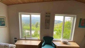 Une pièce dotée de deux fenêtres donnant sur un paysage verdoyant. On y trouve une chaise bleue et une petite table sur laquelle est posé un objet décoratif.