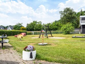 Un parque infantil con columpios y un tobogán, rodeado de césped, un sendero pavimentado y un parterre de flores.