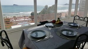 A dining table set with plates, cutlery, and a bowl of fruit, overlooking a beach and ocean view.