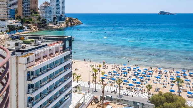 A coastal cityscape with a beach area featuring numerous umbrellas and sun loungers.