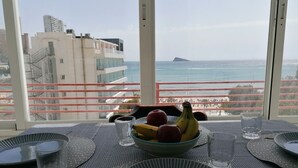A dining table with plates, cutlery, and a bowl of fruit, overlooking a coastal view with buildings and a distant island.