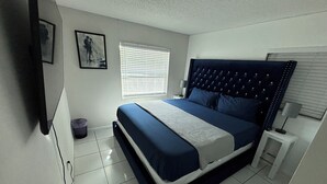 A bedroom with a bed, a window with blinds, a wall-mounted TV, and a trash bin.