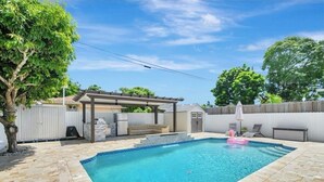 A rectangular swimming pool with a stone tile deck, a pergola, and a small covered seating area.