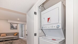 Une buanderie moderne avec un lave-linge et un sèche-linge à chargement frontal, un canapé gris avec un coussin et un tableau encadré accroché au mur.