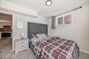 Un dormitorio con cama, mesita de noche, cortinas en la ventana y una cocina visible al fondo.