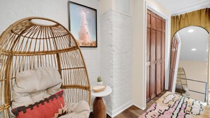 Una habitación con una silla de mimbre, una mesita con un cuenco, un cuadro en la pared y una alfombra estampada en el suelo.