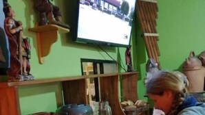 A kitchen area with a person preparing food, a television mounted on the wall, and various kitchen utensils and appliances. - Olas del titicaca backpackers  (Copacabana)