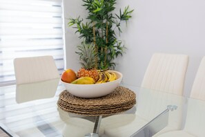 Une table en verre surmontée d’un set de table en fibres tressées, un saladier rempli de fruits et, au fond, une plante en bambou.