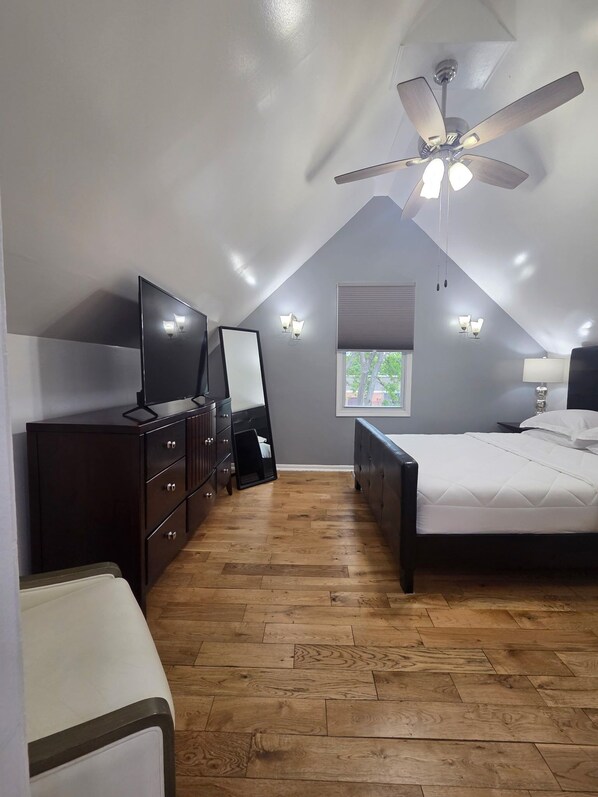 Un dormitorio con techo inclinado, ventilador de techo, cama, cómoda, televisor y dos lámparas fijadas a la pared.