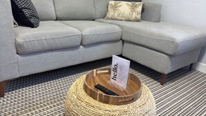 Un sofá gris con un cojín estampado, una cesta tejida con un cartel que dice 'hello' y una alfombra a rayas.