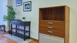 Una habitación con piso de madera, un mueble de madera oscura con cajones, un escritorio pequeño con una planta y dos cuadros en la pared.