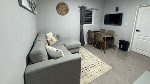 Un salon moderne avec un canapé gris, une table à manger avec des chaises, une horloge au mur et un téléviseur fixé au-dessus de la table.