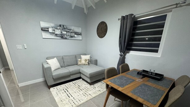 Un salon moderne avec un canapé d’angle gris, une table à manger en bois avec quatre chaises, un tapis à motifs, une horloge murale et une fenêtre avec des rideaux.