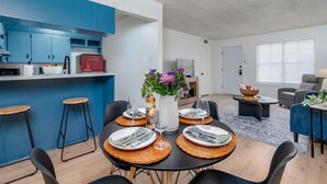 Eine moderne Küche mit blauen Schränken, ein Essbereich mit Tisch für vier Personen und ein Wohnzimmer mit Sofa und Lampe.