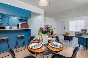A modern kitchen with blue cabinets, a dining area with a table set for four, and a living room with a sofa and a lamp. - Radford Collection Three: Abilene Heights (Abilene)