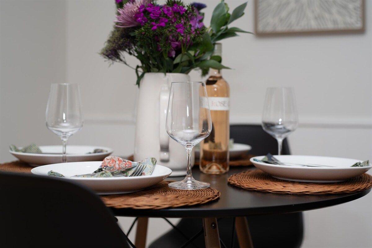 Ein Esstisch mit Tellern, Besteck und Weingläsern, ergänzt durch eine Blumenvase und eine Weinflasche.