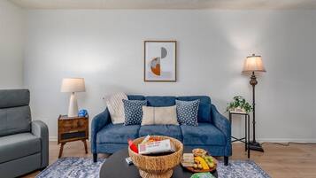 Ein Wohnzimmer mit einem blauen Sofa, einem Couchtisch mit Snacks, einem gerahmten abstrakten Kunstwerk und einer Lampe.