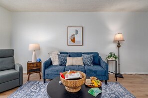 A living room with a blue sofa, a coffee table with snacks, a framed abstract artwork, and a lamp.