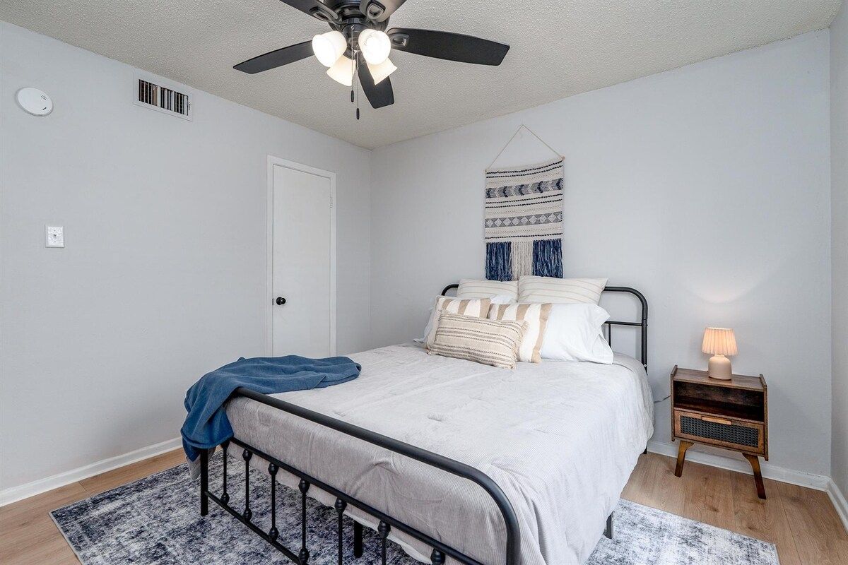 A bedroom with a bed, a ceiling fan, a bedside table with a lamp, and a wall-mounted tapestry.