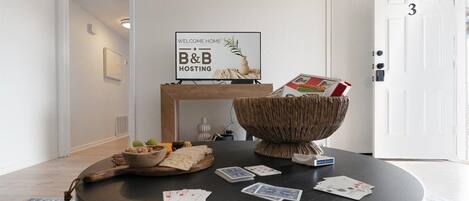 Una mesa redonda con cartas, un cuenco y un cartel de bienvenida en una habitación con una puerta blanca y un cuadro en la pared.
