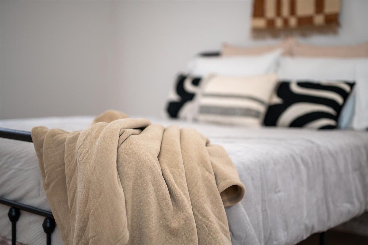 A bed with a beige blanket, a white mattress, and black and white patterned pillows.