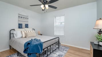 Une chambre à coucher avec un lit, un ventilateur de plafond, une fenêtre avec des stores et une table de chevet avec une lampe.