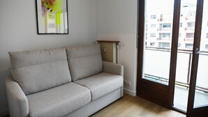 Sofá de tonos neutros en una habitación con radiador, suelo de madera y vista a un balcón con edificios.