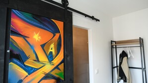 Una habitación con un cuadro abstracto de colores en la pared, un perchero con chaquetas y una puerta que da a otra habitación.