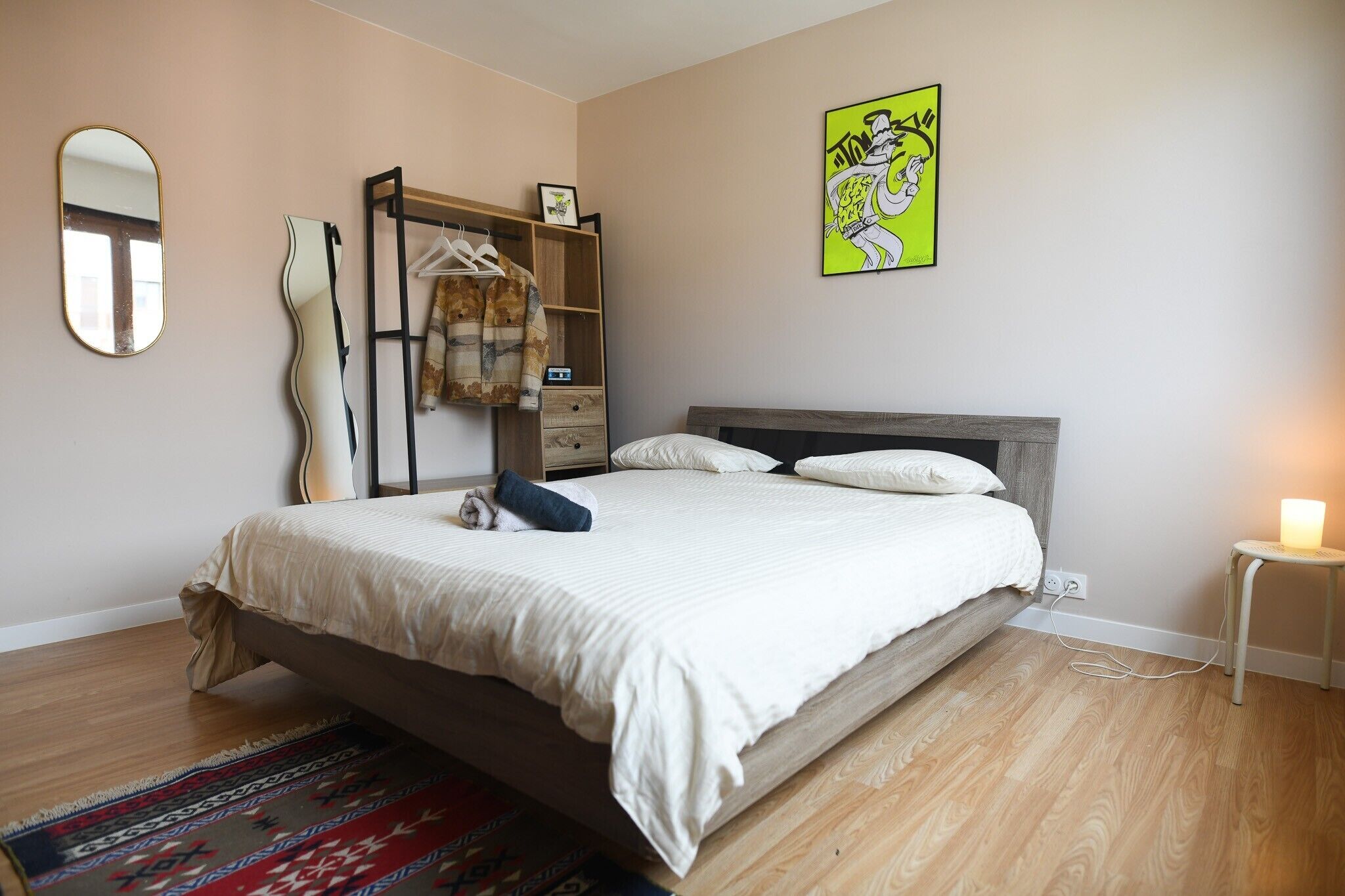 A bedroom with a bed, a bedside table with a lamp, a dresser, a mirror, and a wall art piece.