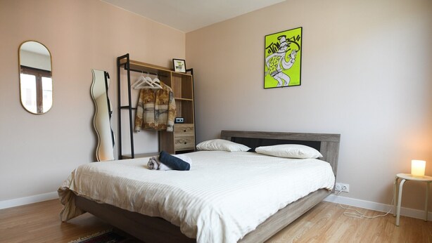 A bedroom with a bed, a bedside table with a lamp, a dresser, a mirror, and a wall art piece.