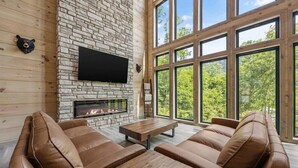 A modern living room with a stone fireplace, leather sofas, a wooden coffee table, and large windows offering a view of the outdoors.