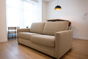 Un sofá color beige en una habitación con suelo de madera, una mesita pequeña, una lámpara colgante y una ventana con cortinas traslúcidas.