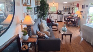 Una sala de estar acogedora con un sofá, una mesa de centro y un televisor. Hay una zona de comedor con una mesa y sillas. El piso es de madera y hay un espejo grande.