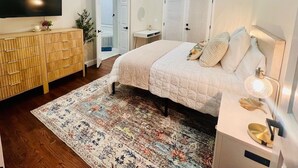 A bedroom with a bed, a TV, a ceiling fan, a dresser, and a door leading to another room.