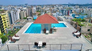 Una piscina sul tetto con un tetto rosso, circondata da poltroncine e tavolini, con vista su una zona urbana densamente edificata.