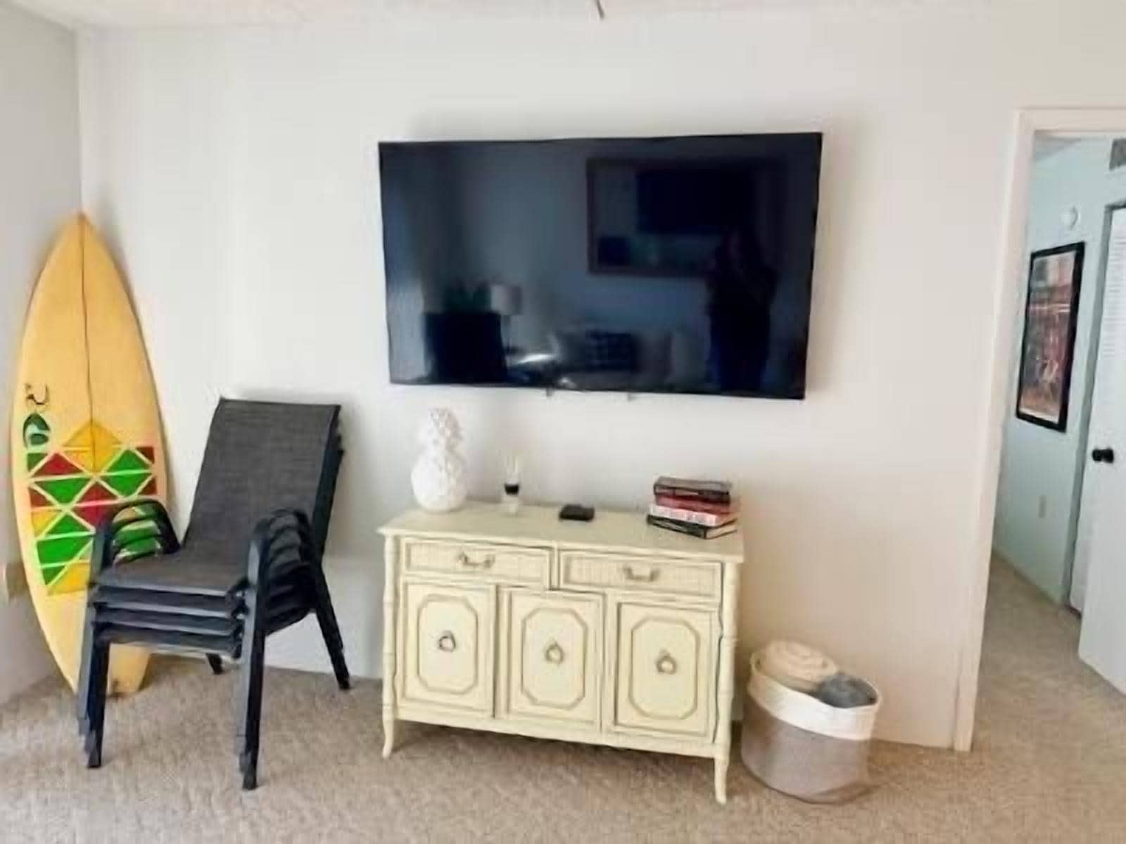 A room with a TV mounted on the wall, a surfboard leaning against the wall, a chair, a small white cabinet, and a door.