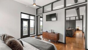 A modern bedroom with a large bed, a dresser, a TV mounted on the wall, and a sliding glass door leading to a balcony.