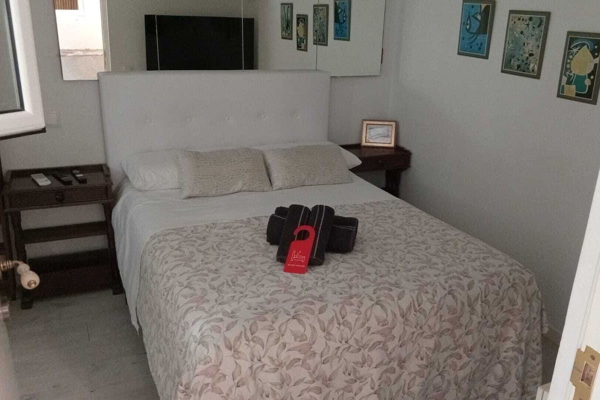 Un dormitorio con una cama, una mesita de noche, un televisor montado en la pared y varias fotografías enmarcadas.