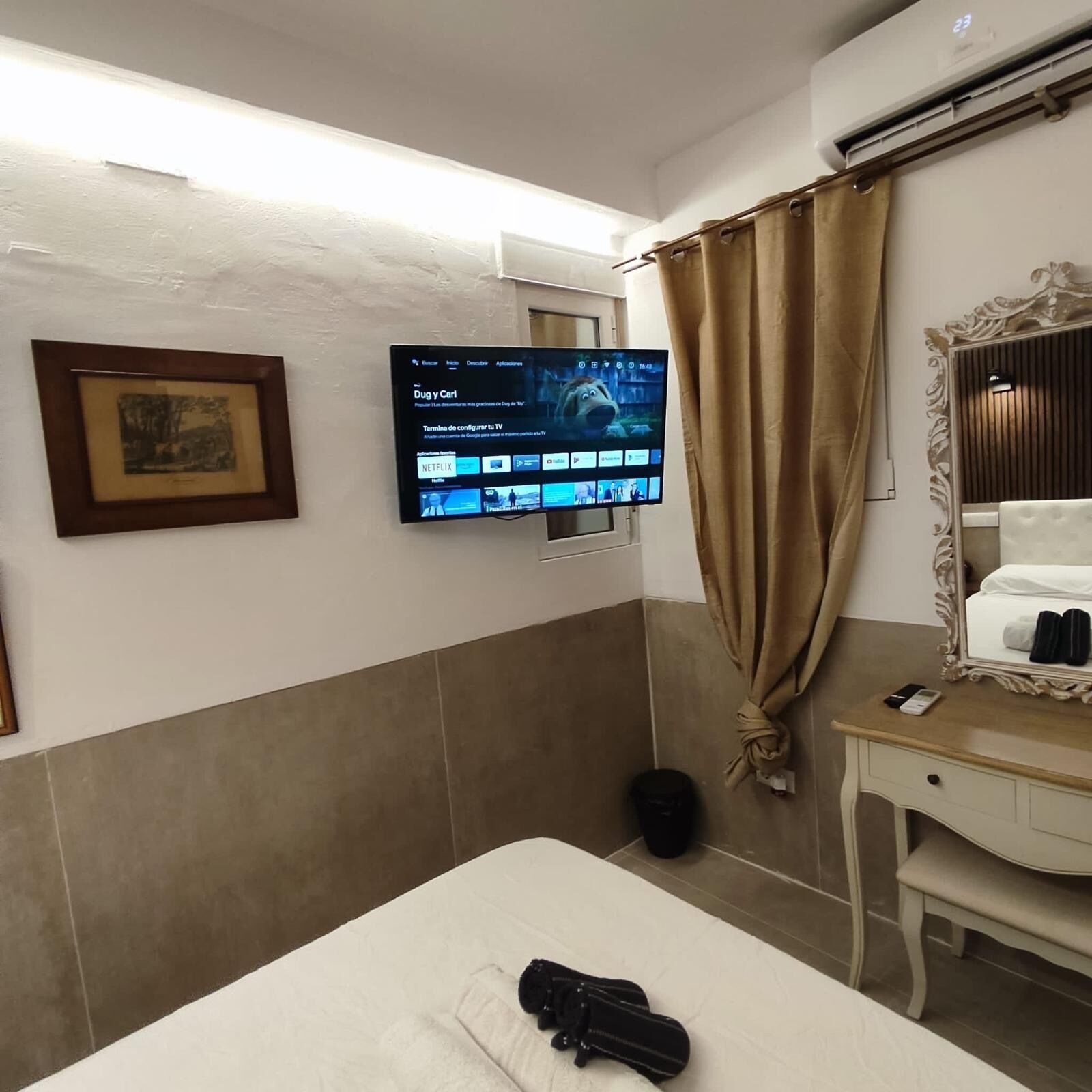A hotel room with a bed, a desk with a mirror, a television mounted on the wall, and a framed picture.