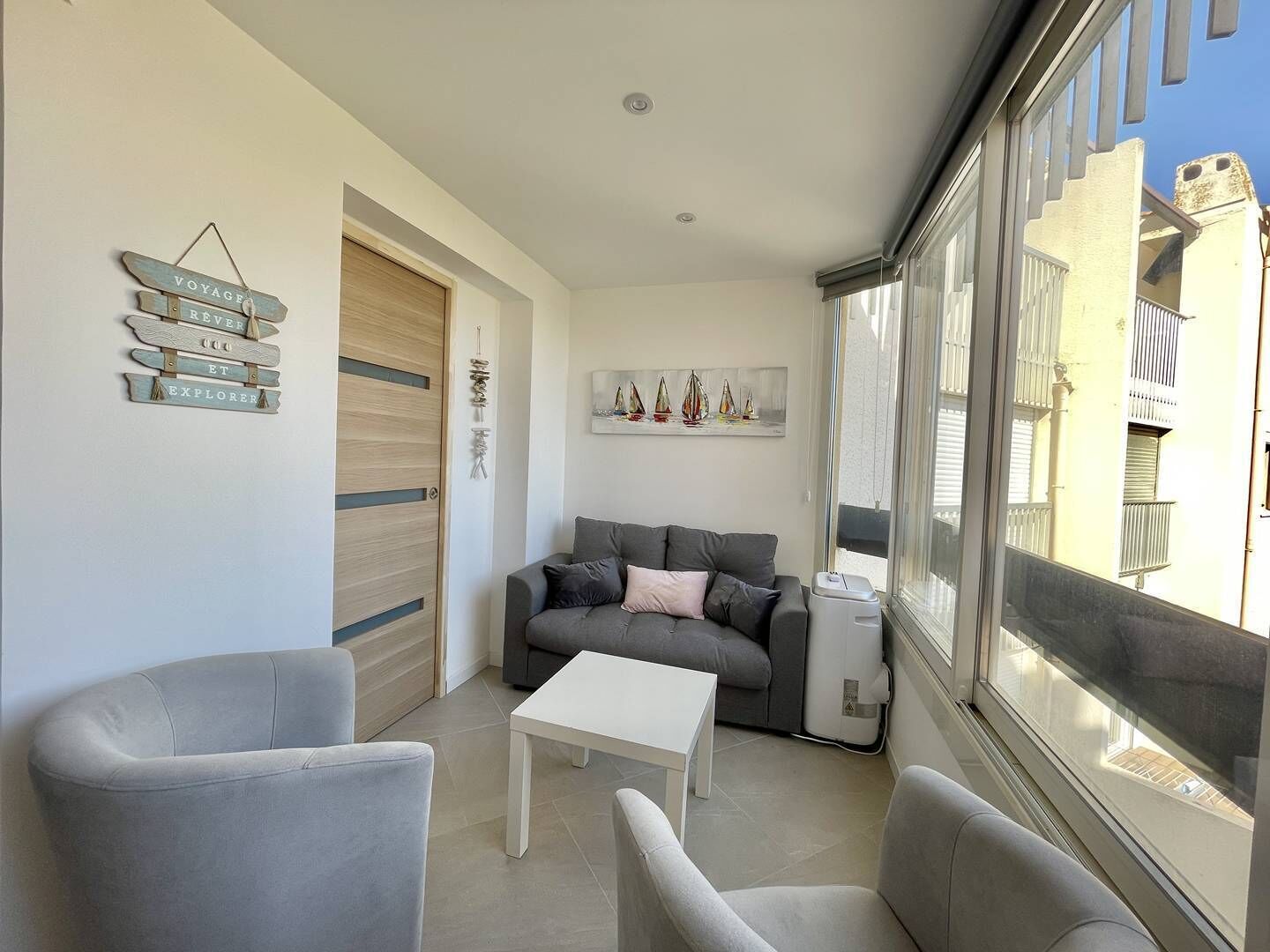 Ein modernes Wohnzimmer mit einem grauen Sofa, zwei Sesseln, einem weißen Couchtisch und einem großen Fenster mit Blick auf Gebäude.