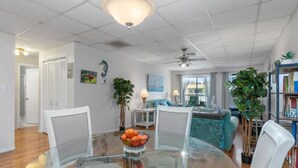 A modern living room with a glass dining table, white chairs, a teal sofa, and a ceiling fan.