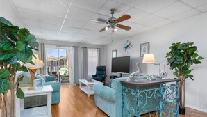 A spacious living area with a sofa, coffee table, and a TV. There are potted plants and a ceiling fan.