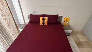 A bed with a red bedspread, two pillows, and a decorative pillow with a logo. There is a nightstand with a lamp and a small table with a yellow object on it. - Peaceful retirement near the station (Marrakech)